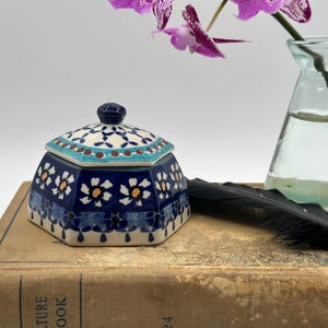 May include: A hexagonal ceramic trinket box with a lid, featuring a blue and white floral pattern. A black feather and a small glass vase with a purple flower are next to the box, resting on a vintage book.