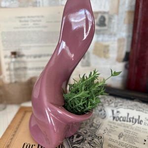 May include: A mauve-colored, abstract ceramic vase with a curved, flowing design. The vase holds a small arrangement of green plants. The vase is placed on a stack of vintage sheet music and books, creating a decorative display.