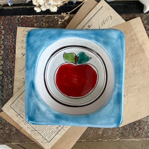 May include: Square ceramic dish with a light blue border and a central design of a red apple with green leaves. The apple is set against a white background with a black border. The dish is likely for decorative use.