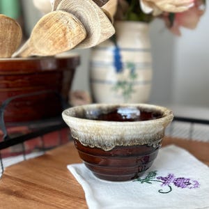 May include: A small, brown ceramic bowl with a cream-colored rim. The bowl has a glossy finish and is sitting on a wooden surface. A white cloth with purple floral embroidery is beneath the bowl. Wooden spoons and a ceramic jar are in the background.