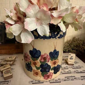 May include: A vintage ceramic crock with a floral design, filled with pink and white faux hydrangeas. The crock has a blue rim and is decorated with blue, pink, and peach flowers. The arrangement sits on sheet music.