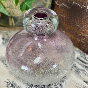 May include: A two-part glass vase with a round, bubble-textured base in a gradient of purple to clear. The top section is a clear, bubble-textured sphere with a deep purple interior. The vase sits on a patterned fabric.