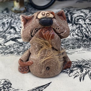 May include: A handmade ceramic figurine of a cat-like creature. The sculpture is primarily brown with a black nose and a feather held in its paws. The figurine has a whimsical, folk art style and is sitting down.