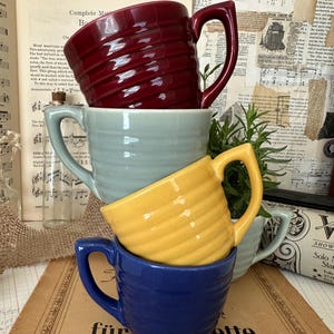 May include: A stack of four ceramic mugs in shades of burgundy, seafoam green, yellow, and blue. Each mug has a ribbed texture and a curved handle. The mugs are stacked on top of each other, with the blue mug at the bottom. The background includes vintage sheet music.