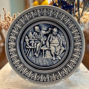 May include: A round, blue and white decorative plate with a detailed relief scene of two figures in a tavern setting. The plate has an ornate border design. The plate is displayed on a clear stand.