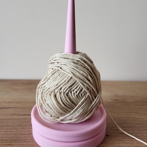 以下が含まれることがあります： ピンク色の糸立てと、薄いベージュ色の糸玉。 糸はピンク色の円錐形のスピンドルに巻き付けられています。 ホルダーは丸いピンク色のベースに置かれています。 糸は自然な色です。