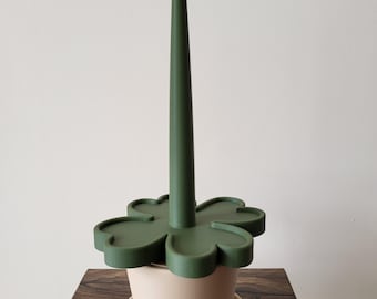 May include: A green, clover-shaped plant watering spike with a tall, pointed center. It sits atop a small, beige ceramic pot with a matching saucer. The items are displayed on a dark wooden surface against a white wall.