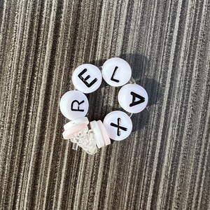 May include: White plastic beads with black letters spelling out the word "RELAX" on a woodgrain background.