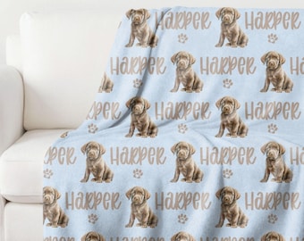 May include: A blue fleece blanket with a repeating pattern of brown puppies and the name "Harper" printed on it. The blanket is folded and displayed on a white couch.