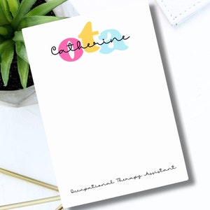 May include: A white notepad with the text "Catherine OTA" in black cursive writing. The letters "OTA" are in colorful circles: pink, yellow, and blue. The text "Occupational Therapy Assistant" is at the bottom of the notepad.