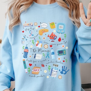 Op de afbeelding: Lichtblauw sweatshirt met een grafisch ontwerp van verschillende medische benodigdheden en de tekst "TID Mama" en "Fight for a cure! Insulin healer".