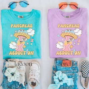 May include: Two light blue and purple t-shirts with a graphic of a cartoon pancreas being abducted by a UFO. The text on the shirts reads "Pancreas Abduction". The shirts are paired with denim shorts and sneakers.