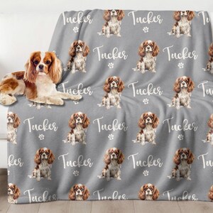May include: A gray fleece blanket with a repeating pattern of a Cavalier King Charles Spaniel dog and the name "Tucker".
