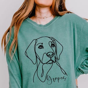 May include: A teal long-sleeve shirt featuring a black line drawing of a dog's face. The word "Grapes" is written in a script font below the dog illustration. The shirt has a relaxed fit and a crew neck.