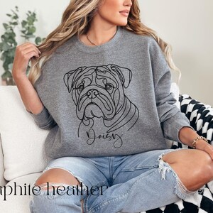K&ouml;nnte beinhalten: Graues Sweatshirt mit Rundhalsausschnitt und einer Strichzeichnung eines Bulldoggen-Gesichts und dem Namen "Daisy" darunter. Das Sweatshirt hat einen l&auml;ssigen Stil. Die Worte "graphite heather" sind unten im Bild aufgedruckt.