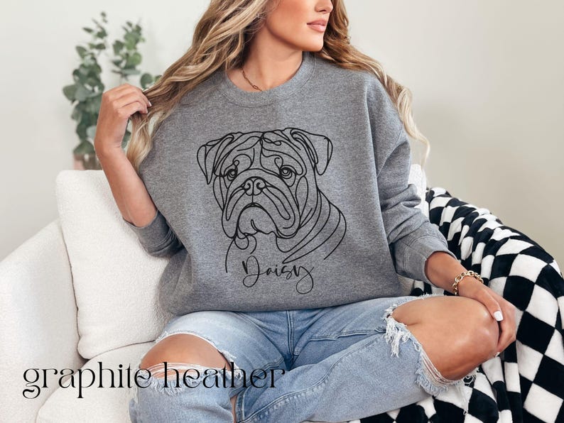 K&ouml;nnte beinhalten: Graues Sweatshirt mit Rundhalsausschnitt und einer Strichzeichnung eines Bulldoggen-Gesichts und dem Namen "Daisy" darunter. Das Sweatshirt hat einen l&auml;ssigen Stil. Die Worte "graphite heather" sind unten im Bild aufgedruckt.