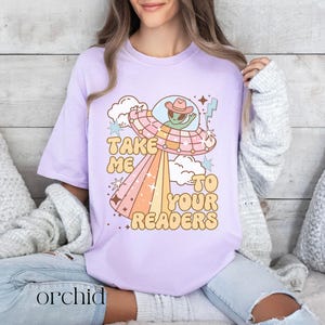 Puede incluir: Camiseta lila claro con un dise&ntilde;o retro de un extraterrestre con sombrero de vaquero en un platillo volante. El texto "TAKE ME TO YOUR READERS" est&aacute; en una fuente divertida. La palabra "orchid" est&aacute; impresa en la parte inferior.