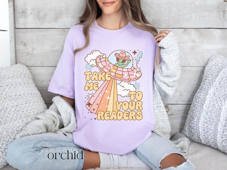 Puede incluir: Camiseta lila claro con un dise&ntilde;o retro de un extraterrestre con sombrero de vaquero en un platillo volante. El texto "TAKE ME TO YOUR READERS" est&aacute; en una fuente divertida. La palabra "orchid" est&aacute; impresa en la parte inferior.