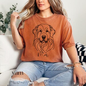 Op de afbeelding: Een vrouw draagt een oranje t-shirt met een zwarte lijntekening van een hondenkop en de naam "Max" erop gedrukt. Ze draagt een blauwe jeans en heeft lang blond haar.