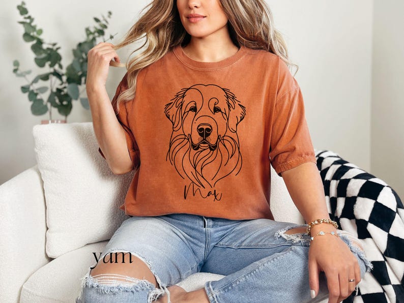 Op de afbeelding: Een vrouw draagt een oranje t-shirt met een zwarte lijntekening van een hondenkop en de naam "Max" erop gedrukt. Ze draagt een blauwe jeans en heeft lang blond haar.