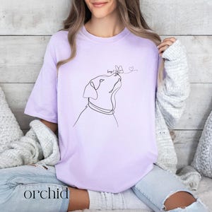 Op de afbeelding: Lichtpaars T-shirt met een lijntekening van een hond die naar een vlinder kijkt. Het woord "boop!" staat boven de vlinder. Het woord "orchid" staat onderaan het shirt.