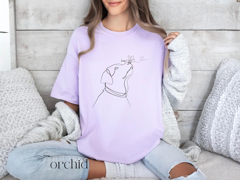 Op de afbeelding: Lichtpaars T-shirt met een lijntekening van een hond die naar een vlinder kijkt. Het woord "boop!" staat boven de vlinder. Het woord "orchid" staat onderaan het shirt.