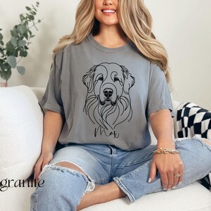 Op de afbeelding: Een grijs T-shirt met een zwarte lijntekening van een hondenkop en de naam "Max" erop gedrukt. De persoon die het shirt draagt, zit op een witte bank.