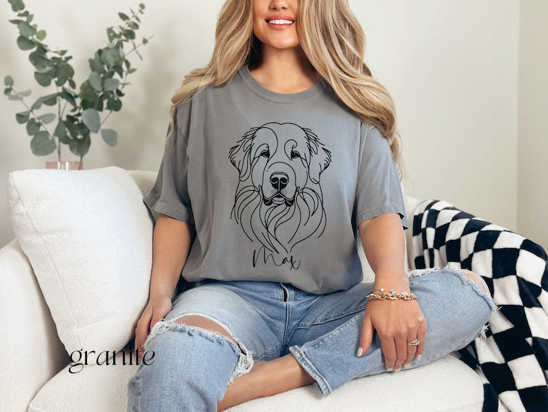 Op de afbeelding: Een grijs T-shirt met een zwarte lijntekening van een hondenkop en de naam "Max" erop gedrukt. De persoon die het shirt draagt, zit op een witte bank.