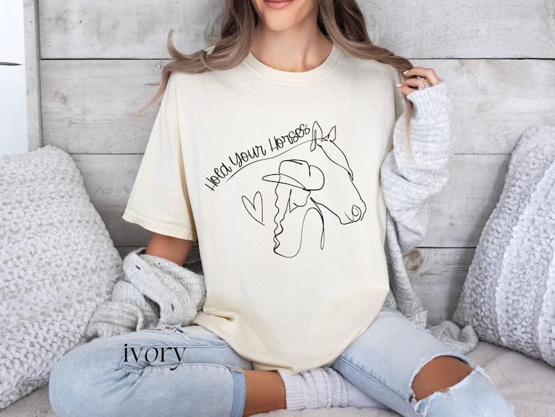 Op de afbeelding: Een vrouw draagt een lichtbeige T-shirt met een zwarte lijntekening van een vrouw die een cowboyhoed en een paard draagt. De tekst "Hold your horses" staat in een gebogen lijn boven de tekening.