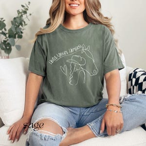 Op de afbeelding: Een vrouw draagt een saliegroen T-shirt met de tekst "Hold your horses" en een lijntekening van een vrouw met een paard. Ze zit op een witte bank met een zwart-wit geblokte deken.