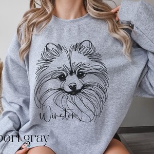 May include: A light gray sweatshirt featuring a black line drawing of a Pomeranian dog's face. The dog's name, "Winston," is written in cursive below the image. The word "sport gray" is printed at the bottom of the sweatshirt.