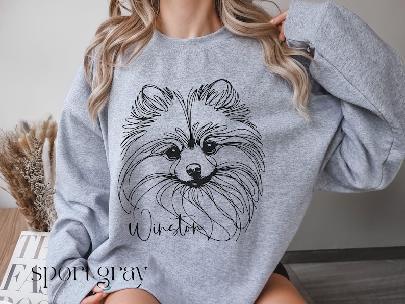 May include: A light gray sweatshirt featuring a black line drawing of a Pomeranian dog's face. The dog's name, "Winston," is written in cursive below the image. The word "sport gray" is printed at the bottom of the sweatshirt.