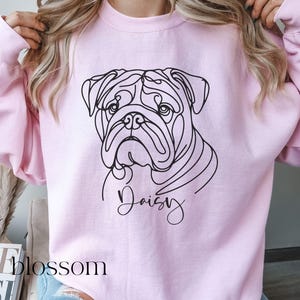 K&ouml;nnte beinhalten: Hellrosa Sweatshirt mit einer schwarzen Strichzeichnung eines Bulldoggen-Gesichts. Der Name des Hundes, "Daisy", ist in Kursivschrift unter dem Bild geschrieben. Das Wort "blossom" ist in der linken unteren Ecke sichtbar.