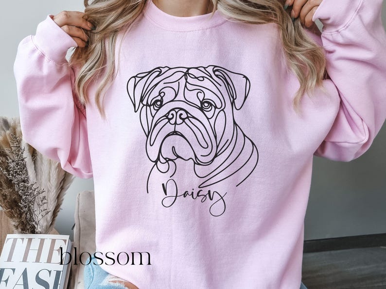 K&ouml;nnte beinhalten: Hellrosa Sweatshirt mit einer schwarzen Strichzeichnung eines Bulldoggen-Gesichts. Der Name des Hundes, "Daisy", ist in Kursivschrift unter dem Bild geschrieben. Das Wort "blossom" ist in der linken unteren Ecke sichtbar.