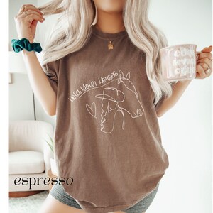 Op de afbeelding: Een bruin T-shirt met de tekst "Hold Your Horses" en een lijntekening van een vrouw die paardrijdt.