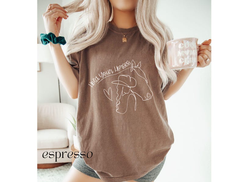 Op de afbeelding: Een bruin T-shirt met de tekst "Hold Your Horses" en een lijntekening van een vrouw die paardrijdt.