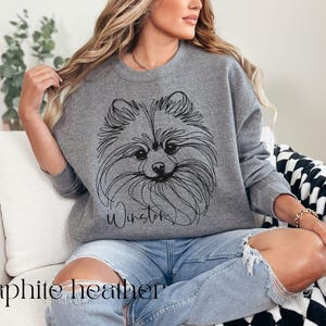 May include: Graphite heather sweatshirt featuring a line drawing of a Pomeranian dog with the name "Winston" in script. The text "graphite heather" is at the bottom. The sweatshirt is worn with blue jeans.