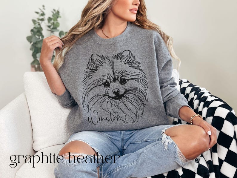 May include: Graphite heather sweatshirt featuring a line drawing of a Pomeranian dog with the name "Winston" in script. The text "graphite heather" is at the bottom. The sweatshirt is worn with blue jeans.