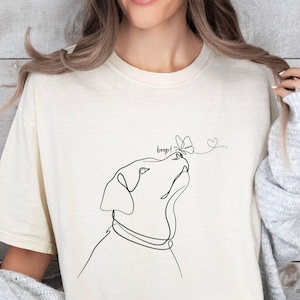 Op de afbeelding: Cr&egrave;mekleurig T-shirt met een minimalistische lijntekening van een hond die naar een vlinder kijkt. Het woord "beep!" staat boven de neus van de hond, met een stippellijn die naar een hart leidt.