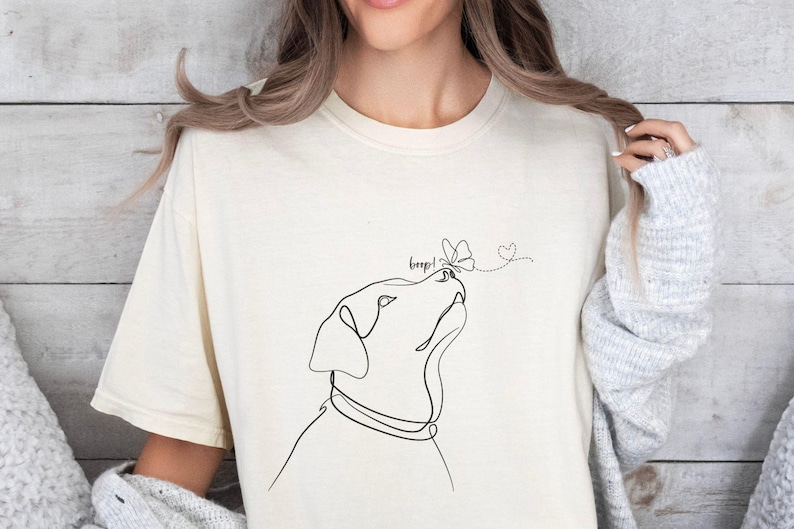 Op de afbeelding: Cr&egrave;mekleurig T-shirt met een minimalistische lijntekening van een hond die naar een vlinder kijkt. Het woord "beep!" staat boven de neus van de hond, met een stippellijn die naar een hart leidt.