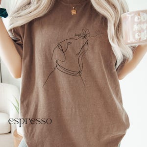 Op de afbeelding: Espresso-kleurig T-shirt met een minimalistische lijntekening van een hond die naar een vlinder kijkt. Het woord "beep!" staat boven de neus van de hond. Het woord "espresso" staat onderaan het shirt.