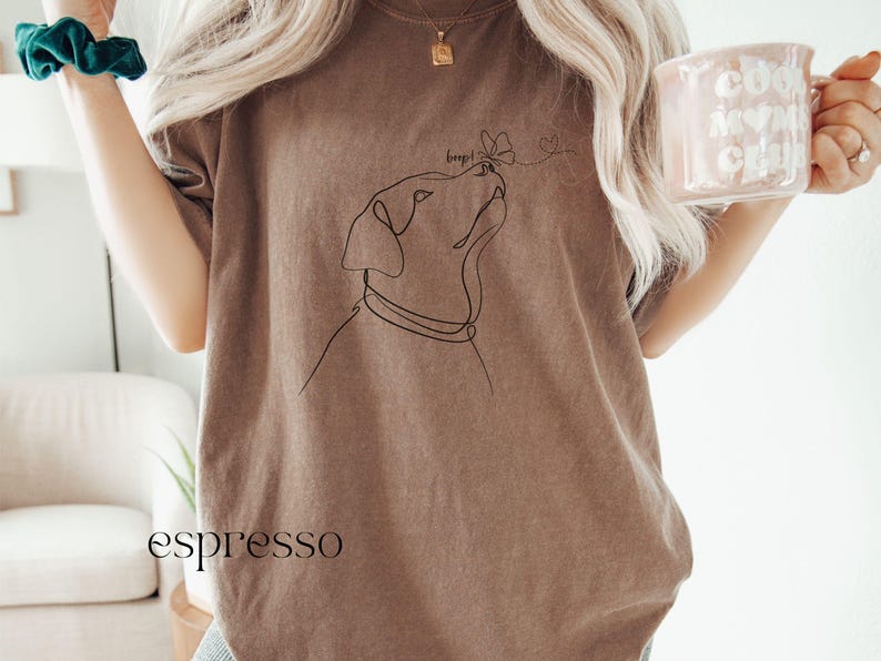 Op de afbeelding: Espresso-kleurig T-shirt met een minimalistische lijntekening van een hond die naar een vlinder kijkt. Het woord "beep!" staat boven de neus van de hond. Het woord "espresso" staat onderaan het shirt.