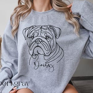 K&ouml;nnte beinhalten: Hellgraues Sweatshirt mit einer schwarzen Strichzeichnung eines Bulldoggen-Gesichts und dem Namen "Daisy" in Schreibschrift. Die Worte "sport gray" sind unten auf dem Sweatshirt aufgedruckt.