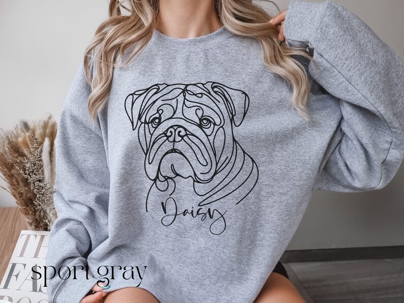 K&ouml;nnte beinhalten: Hellgraues Sweatshirt mit einer schwarzen Strichzeichnung eines Bulldoggen-Gesichts und dem Namen "Daisy" in Schreibschrift. Die Worte "sport gray" sind unten auf dem Sweatshirt aufgedruckt.