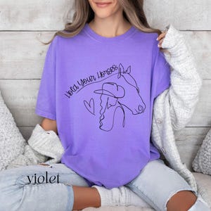 Op de afbeelding: Een paars T-shirt met een zwarte lijntekening van een vrouw die een cowboyhoed en een paard draagt. De tekst "Hold Your Horses" is geschreven in een cursief schrift.