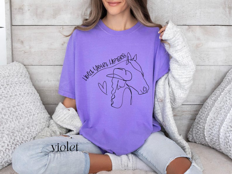 Op de afbeelding: Een paars T-shirt met een zwarte lijntekening van een vrouw die een cowboyhoed en een paard draagt. De tekst "Hold Your Horses" is geschreven in een cursief schrift.