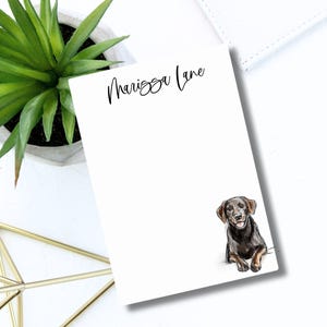 May include: White notepad with the name "Marissa Lane" in elegant script. A watercolor illustration of a black and brown dog is in the lower right corner. A potted succulent and a gold geometric object are in the background.