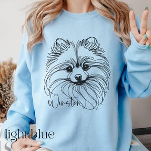 May include: Light blue sweatshirt featuring a black line drawing of a Pomeranian dog's face. The dog's name, "Winston," is written in cursive below the image. The words "light blue" are printed at the bottom of the image.