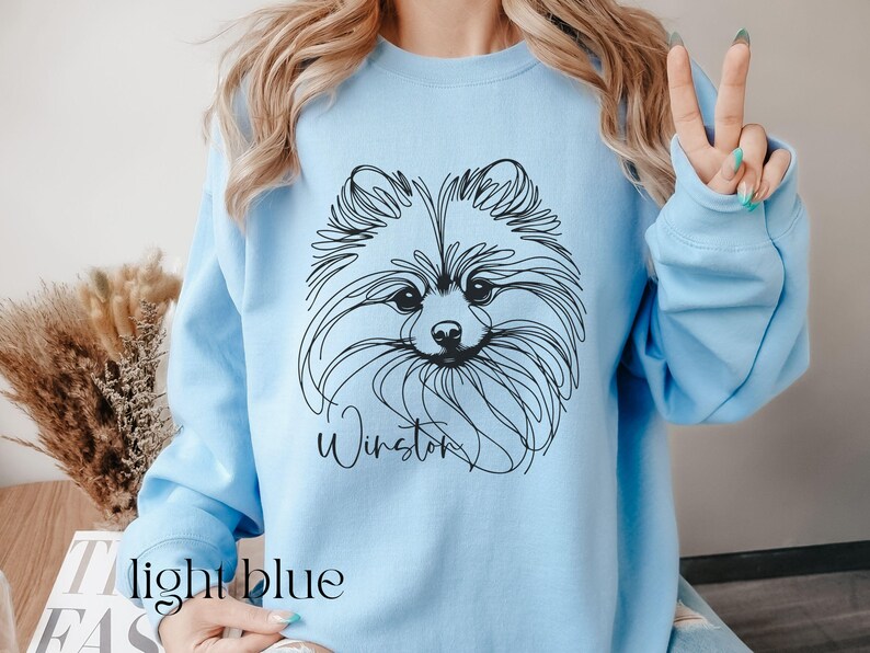 May include: Light blue sweatshirt featuring a black line drawing of a Pomeranian dog's face. The dog's name, "Winston," is written in cursive below the image. The words "light blue" are printed at the bottom of the image.