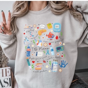 Op de afbeelding: Een lichtgrijs sweatshirt met een kleurrijk grafisch ontwerp met een pancreas, een insulinepomp, een bloedglucosemeter en andere diabetesgerelateerde afbeeldingen. De tekst op het sweatshirt luidt "My kid is a warrior", "T1D Mama", "Love never gives up", "Fight for a cure", "Insulin dealer" en "Snack packer".